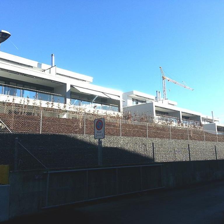 Moderne Wohngebäude auf einem Hang hinter Zaun und Mauer bei sonnigem Himmel