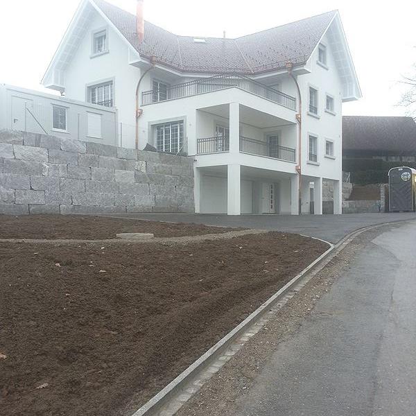 Weißes Wohnhaus mit Satteldach an einer Straße, davor ein Hang mit Erde und eine Steinmauer